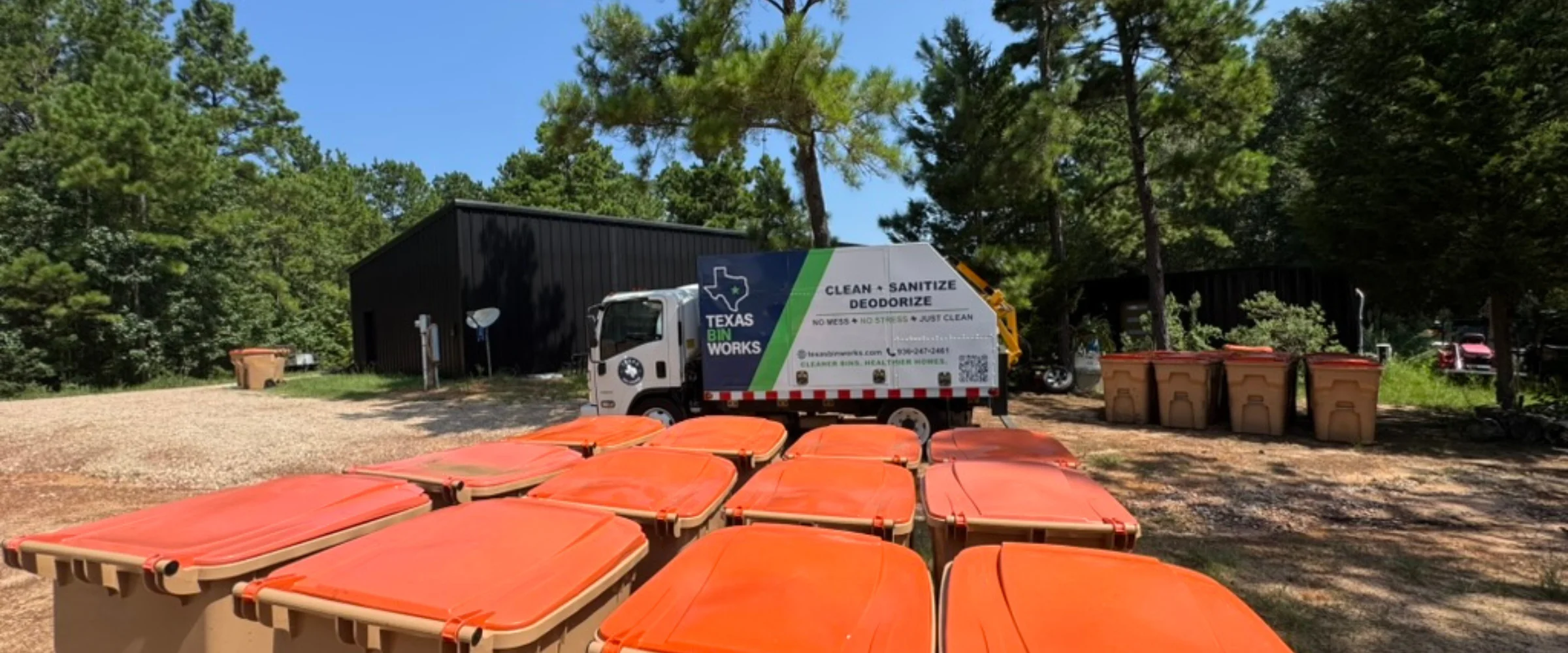A white Texas Bin Works truck with green and blue branding cleans a residential trash can on a sunny street. A white Texas Bin Works truck with green and blue branding cleans a residential trash can on a sunny street.