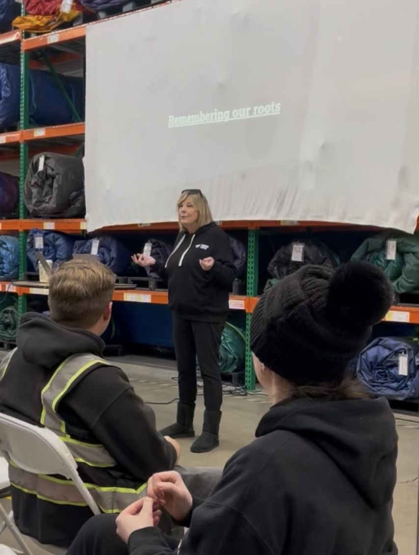 Joy speaking to employees in the warehouse beneath a presentation titled Remembering our roots