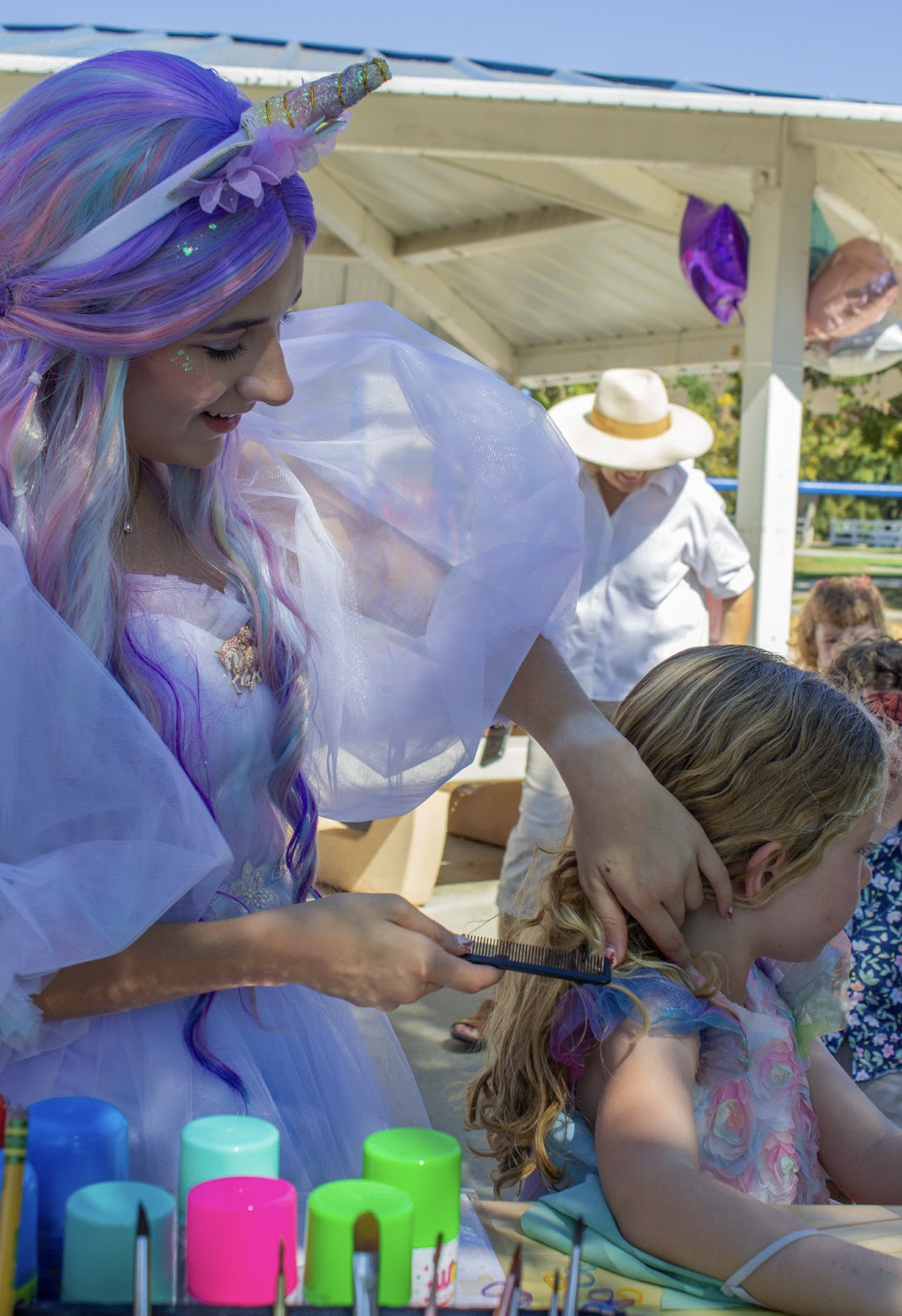 A unicorn-themed performer adding colorful details to a young guest's hair at an outdoor party station