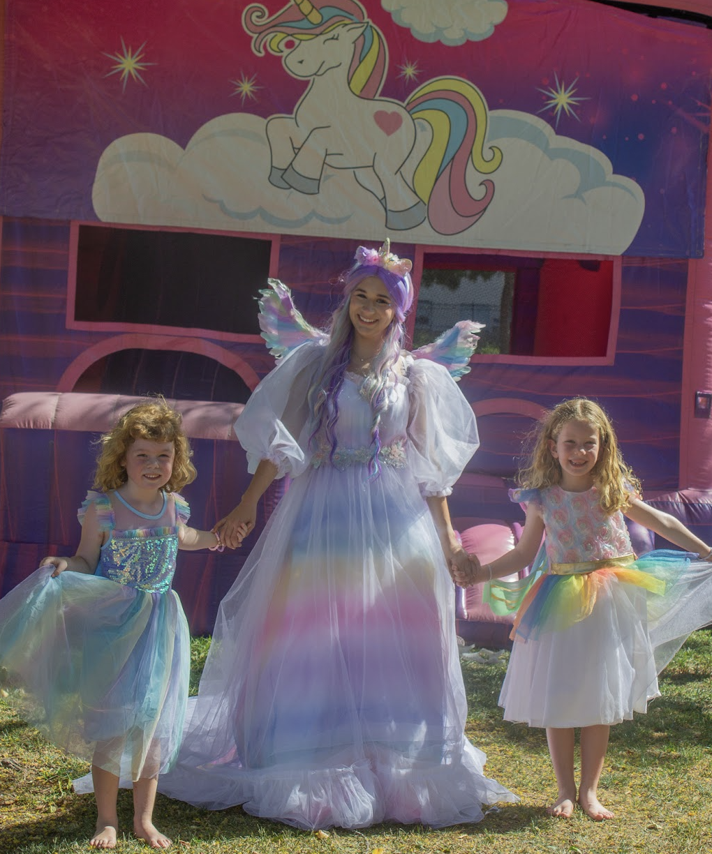 A unicorn-themed character posing with two children beside a matching unicorn bounce house