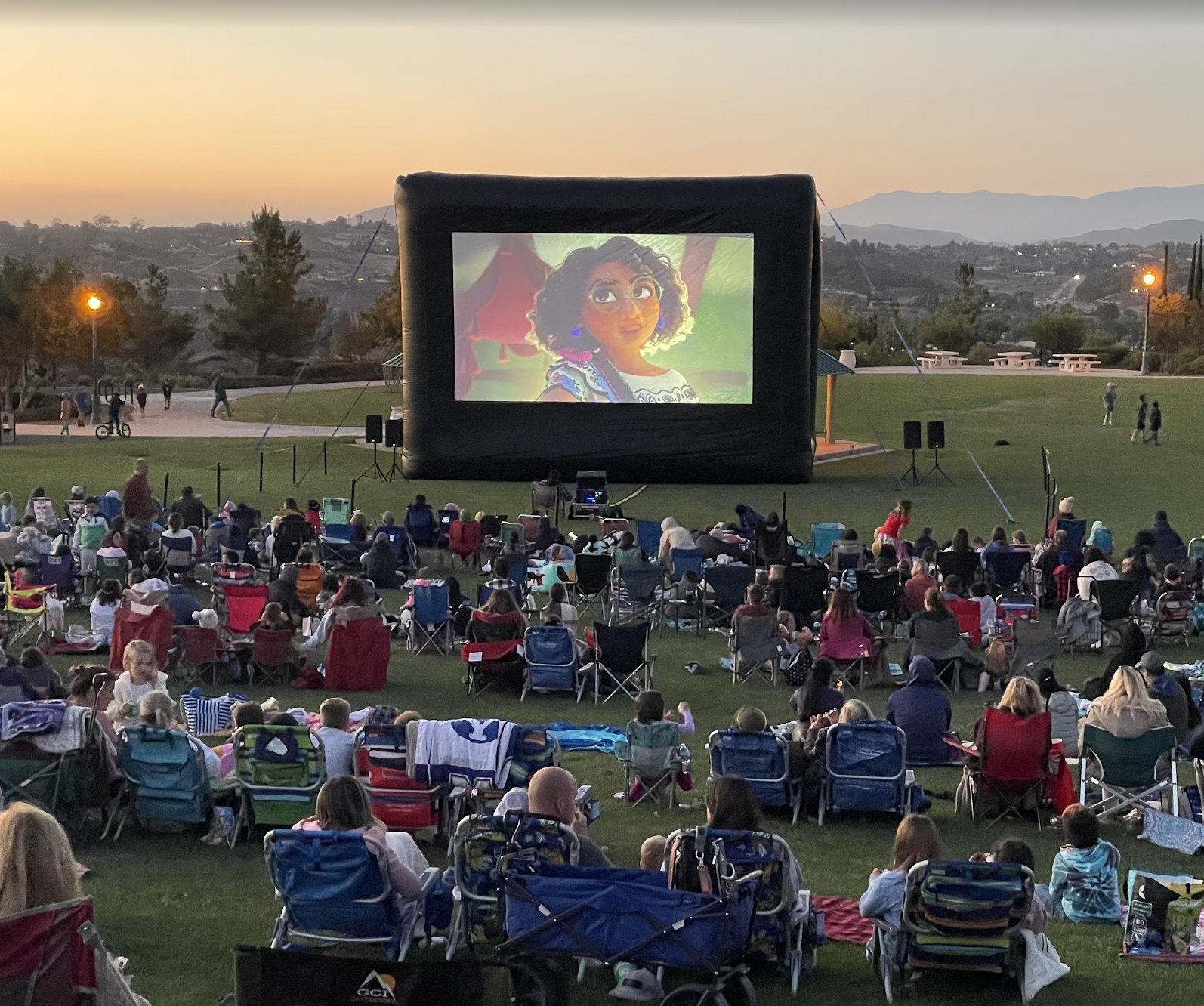 Families seated on a grassy hillside watching an outdoor movie on an inflatable screen at sunset