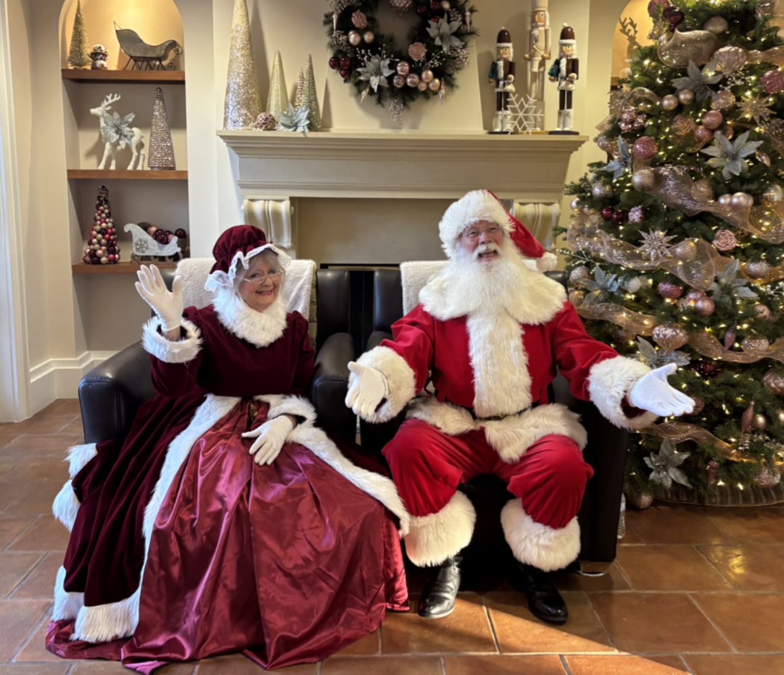 Santa Claus and Mrs. Claus seated beside a decorated Christmas tree and festive fireplace