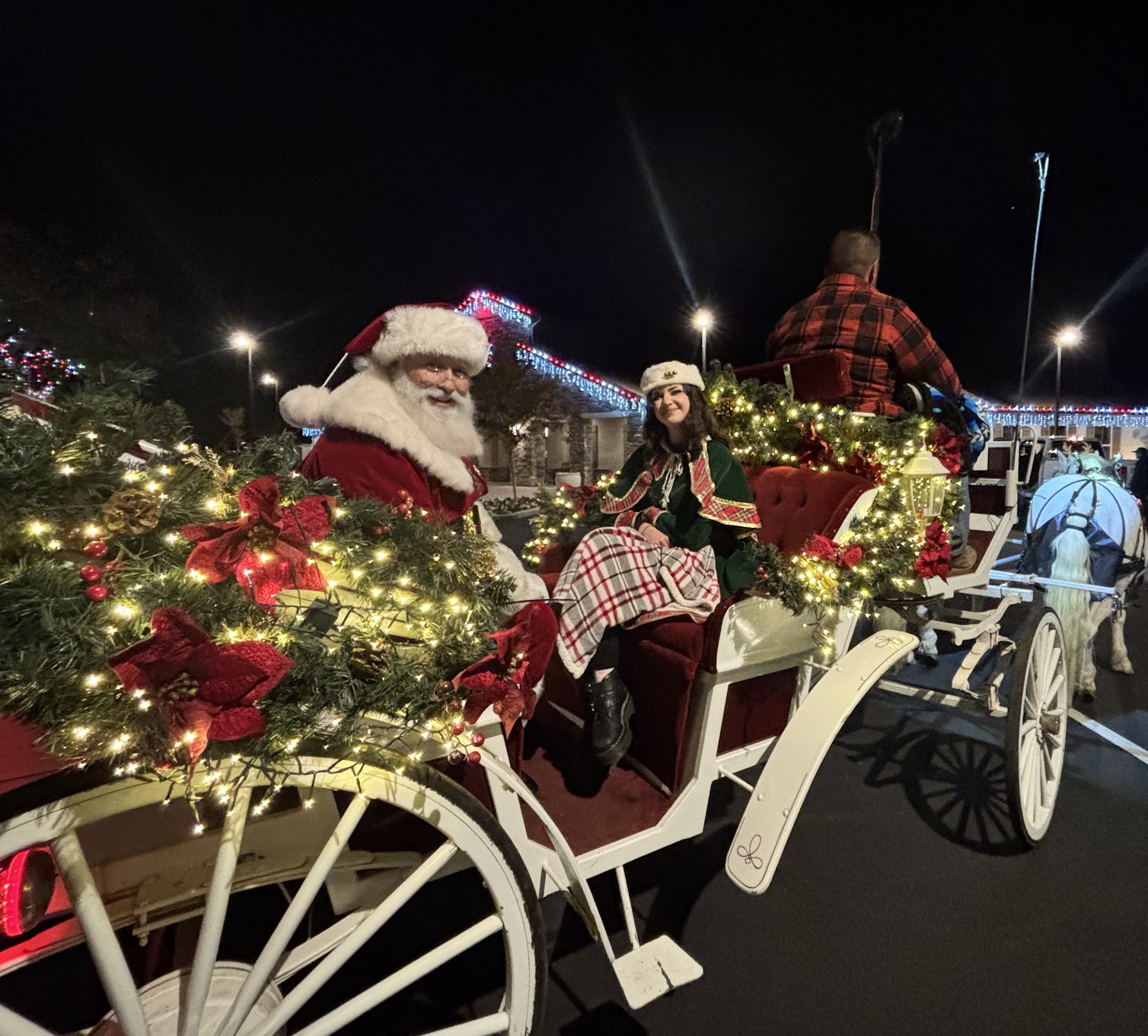 Santa riding in a white horse-drawn carriage decorated with holiday lights and greenery at an evening event
