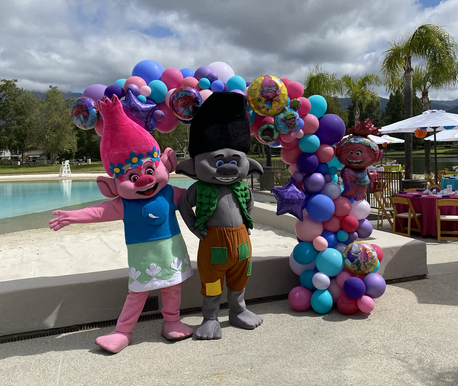 Two colorful troll mascots posing beneath a balloon arch at an outdoor poolside event
