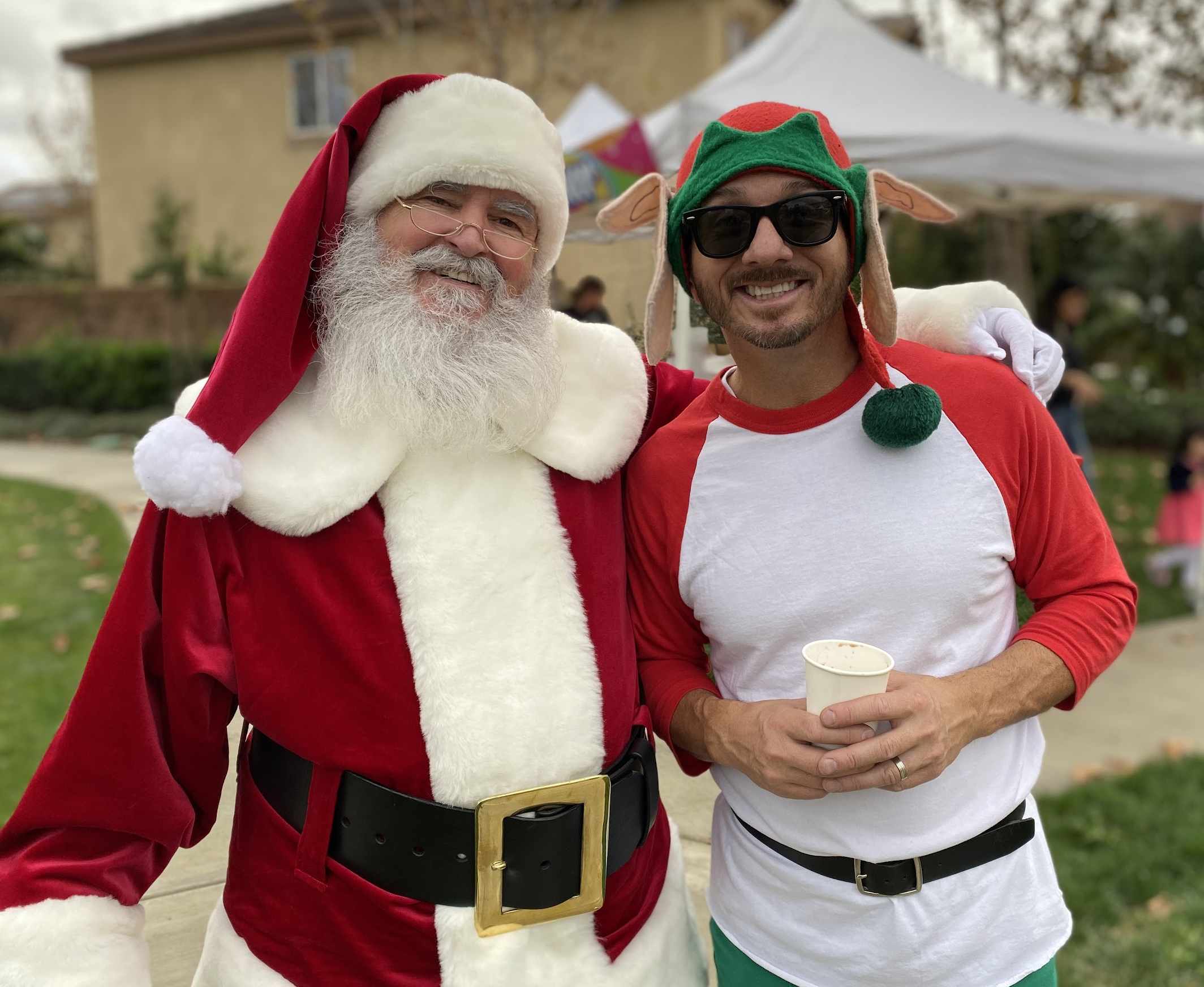 Santa smiling with an adult guest wearing an elf-themed hat and holiday shirt at an outdoor event