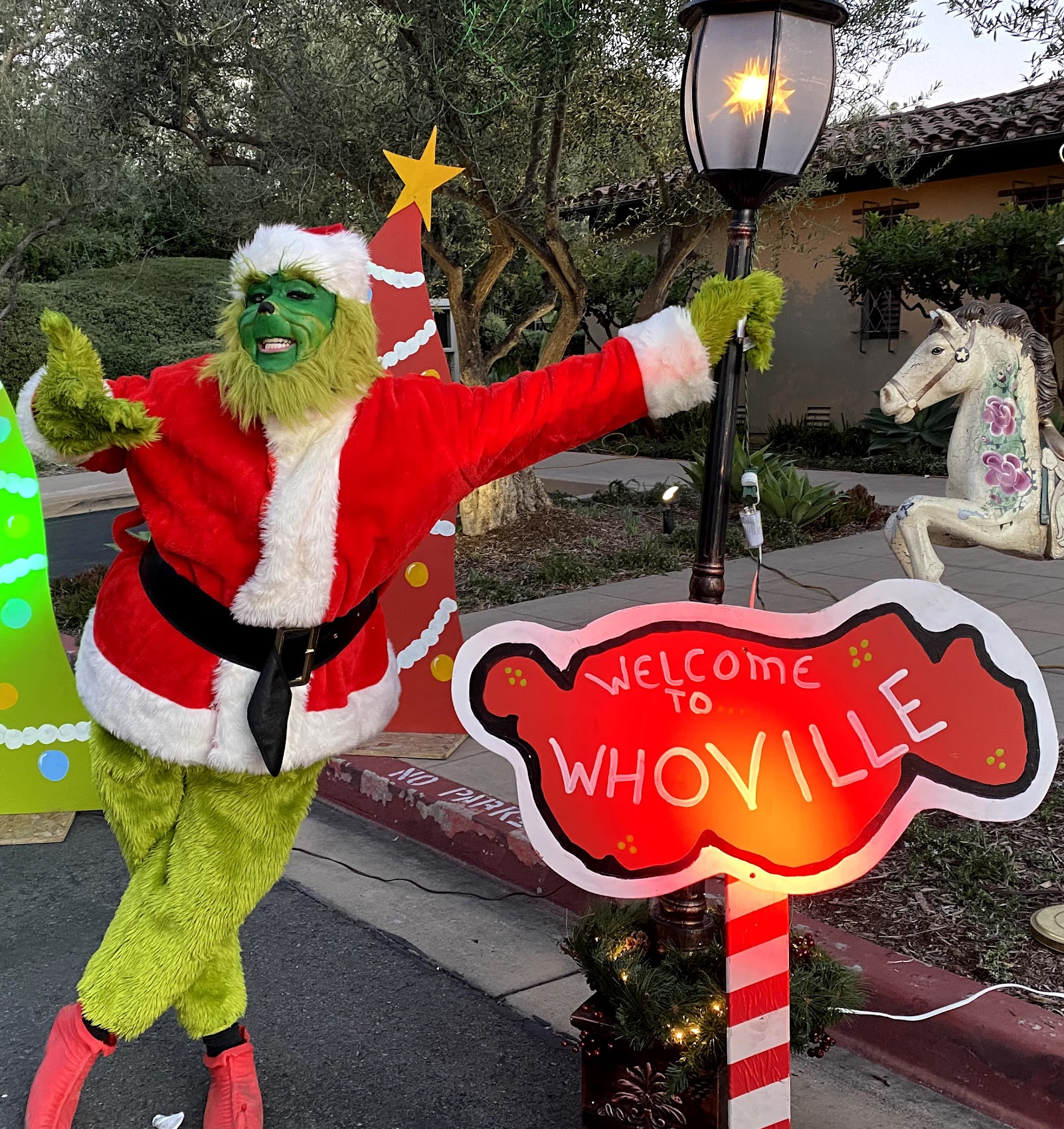 A green holiday character posing beside a Welcome to Whoville sign and festive props