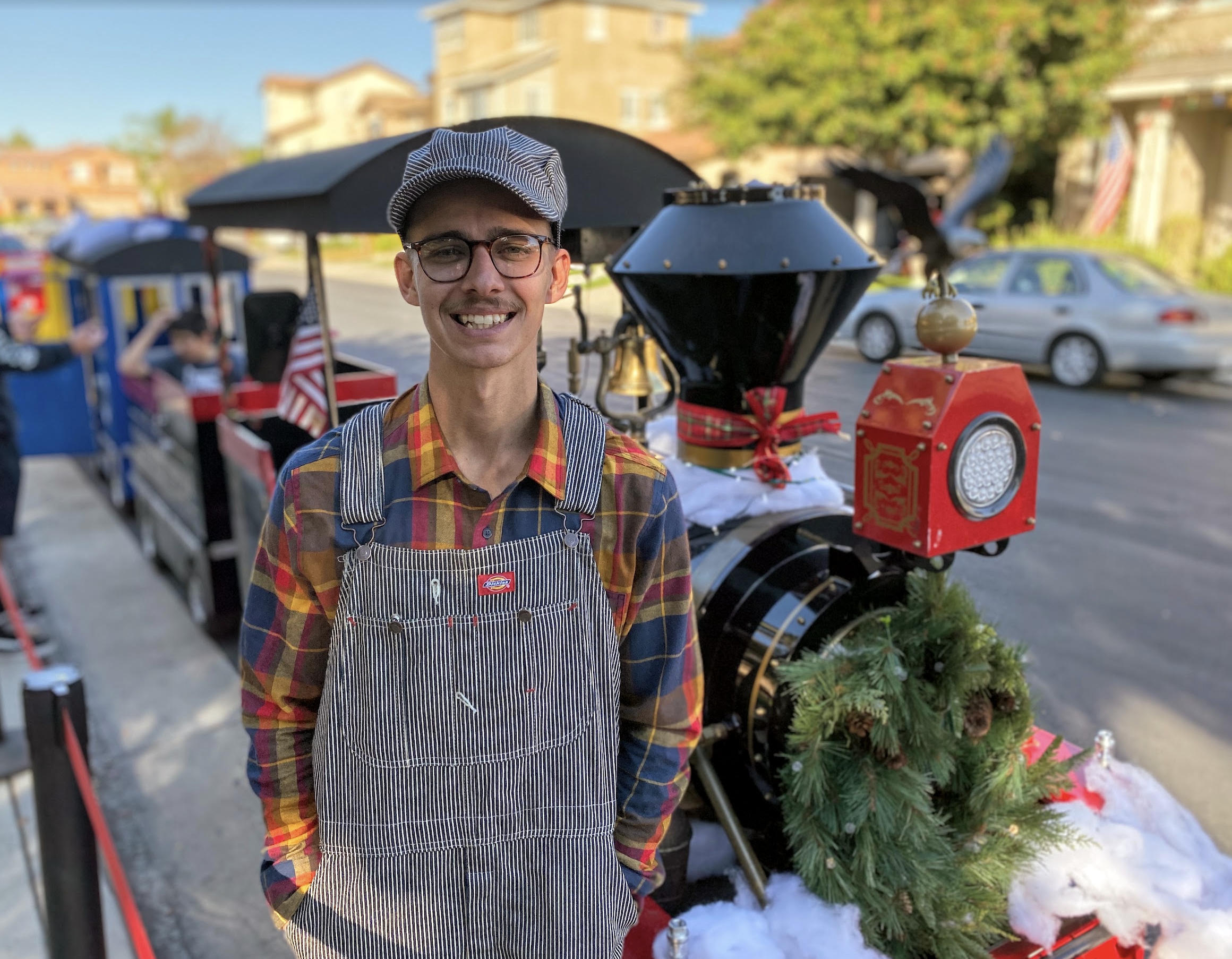 Jolly Express Trackless Train engineer beside a holiday-decorated locomotive with greenery, ribbon and snowy accents