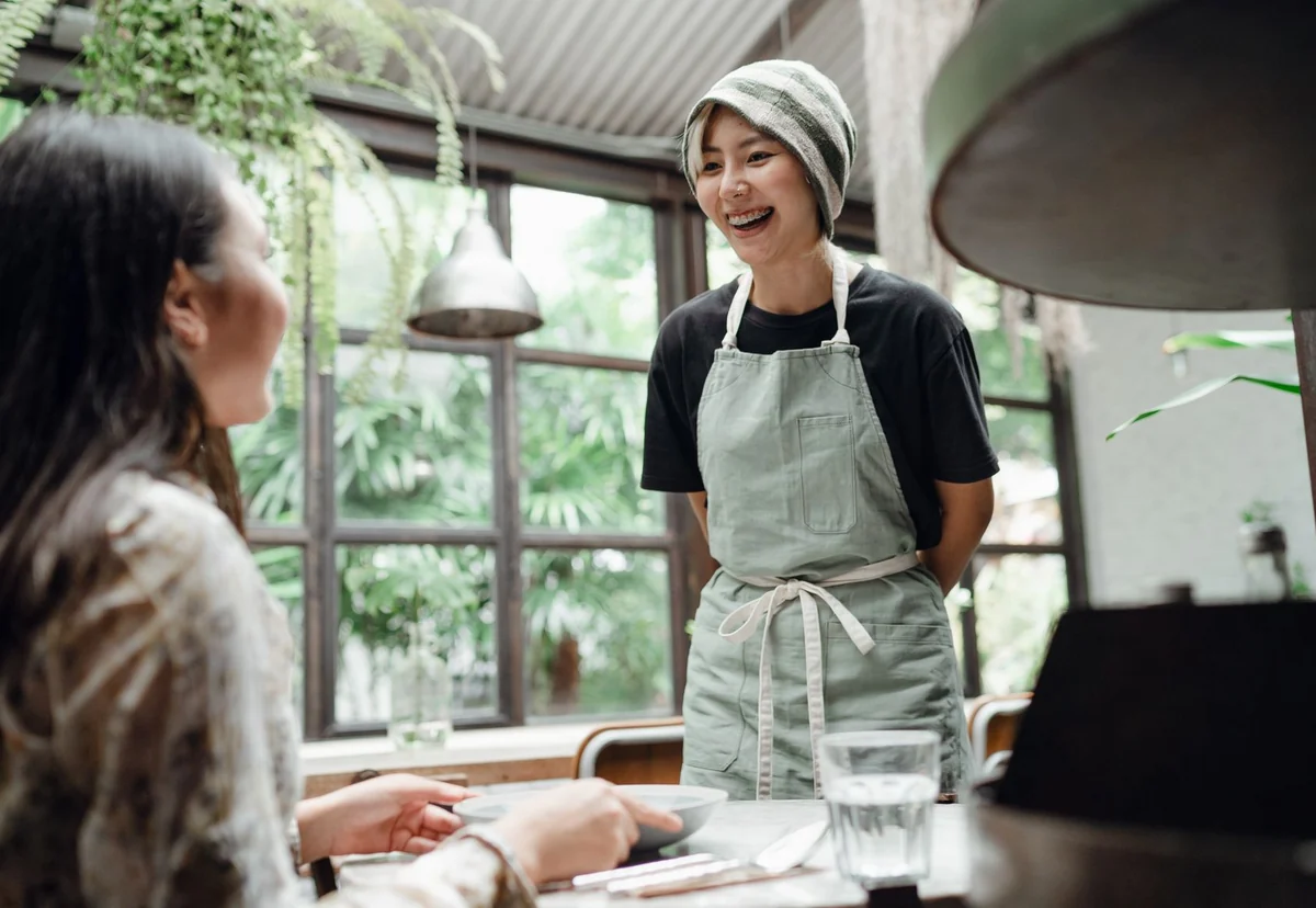 A smiling cafe server in an apron chatting with a customer at her table
