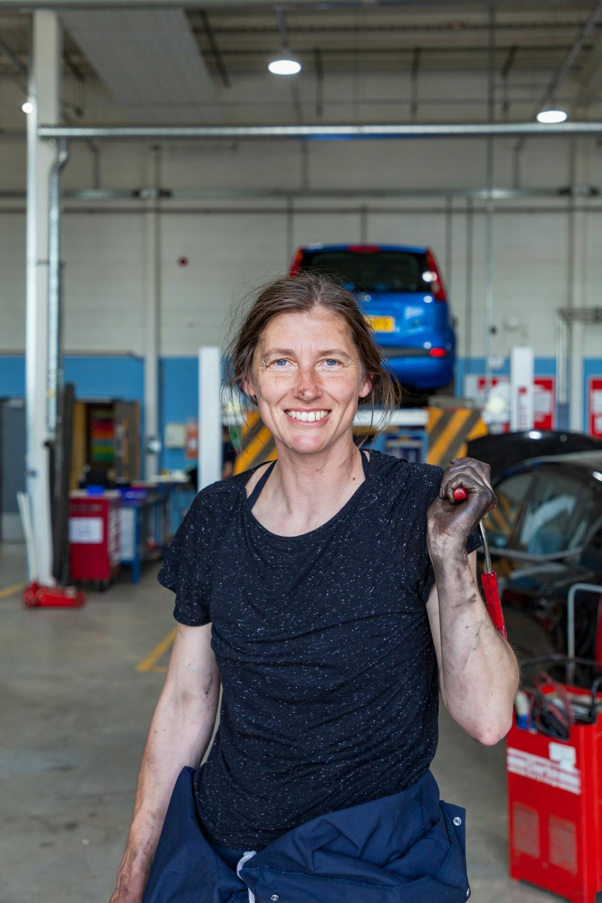 A smiling woman holding a tool in a vehicle workshop