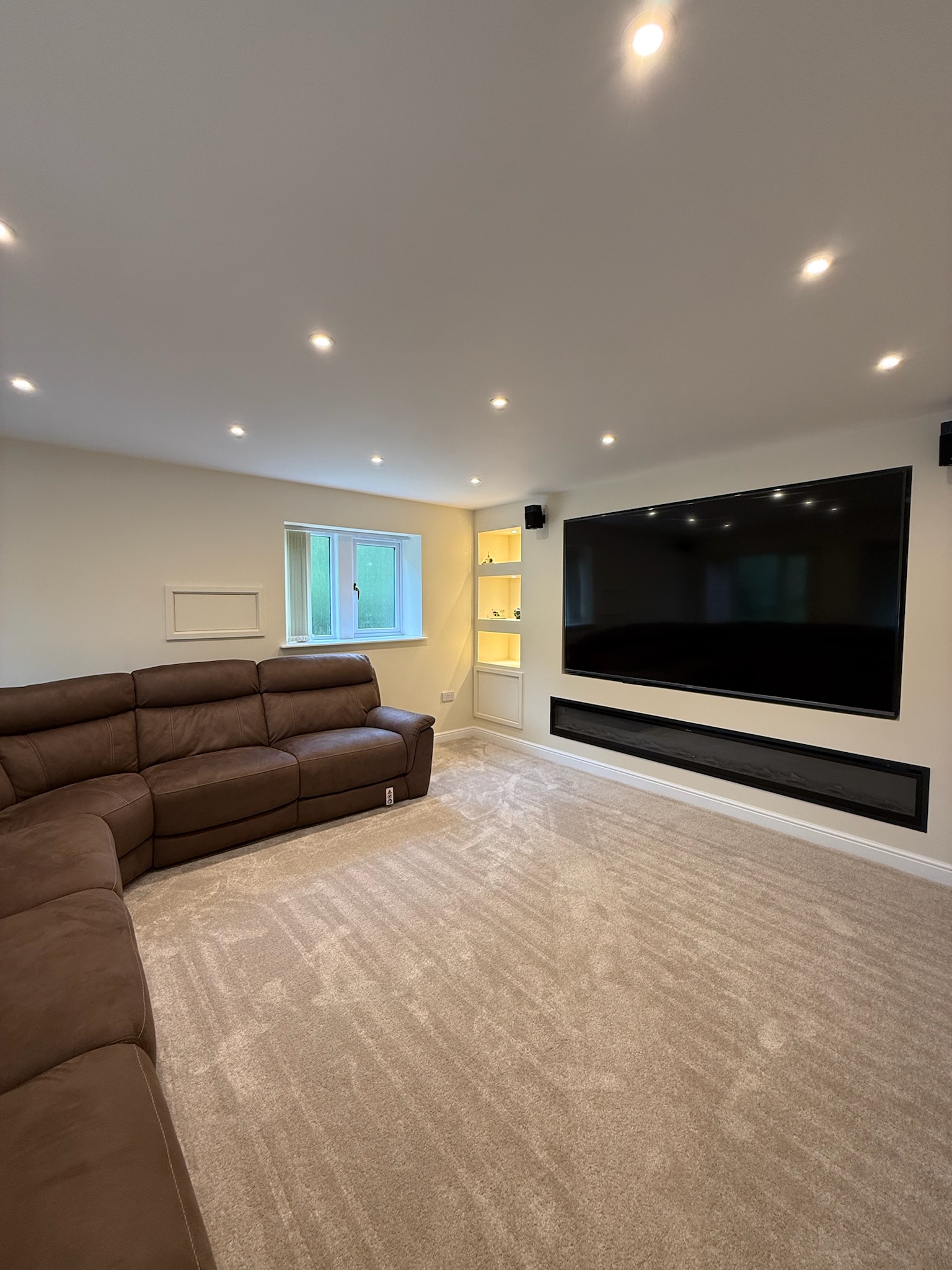 Interior of a completed double garage conversion in Rochdale transformed into a spacious family living room with fitted media wall, recessed lighting, soft carpeting and a large corner sofa, creating additional living space for the home.