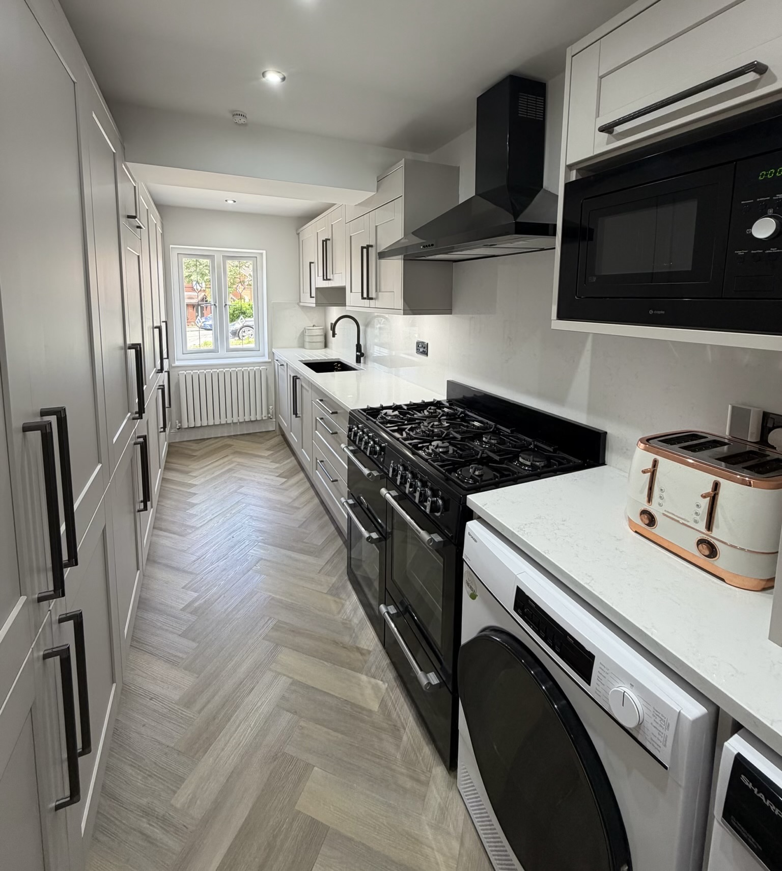 Garage conversion in Ashton transformed into a modern kitchen with fitted cupboards, integrated appliances, herringbone flooring, radiator and a window providing natural light. Garage conversion in Ashton transformed into a modern kitchen with fitted cupboards, integrated appliances, herringbone flooring, radiator and a window providing natural light.