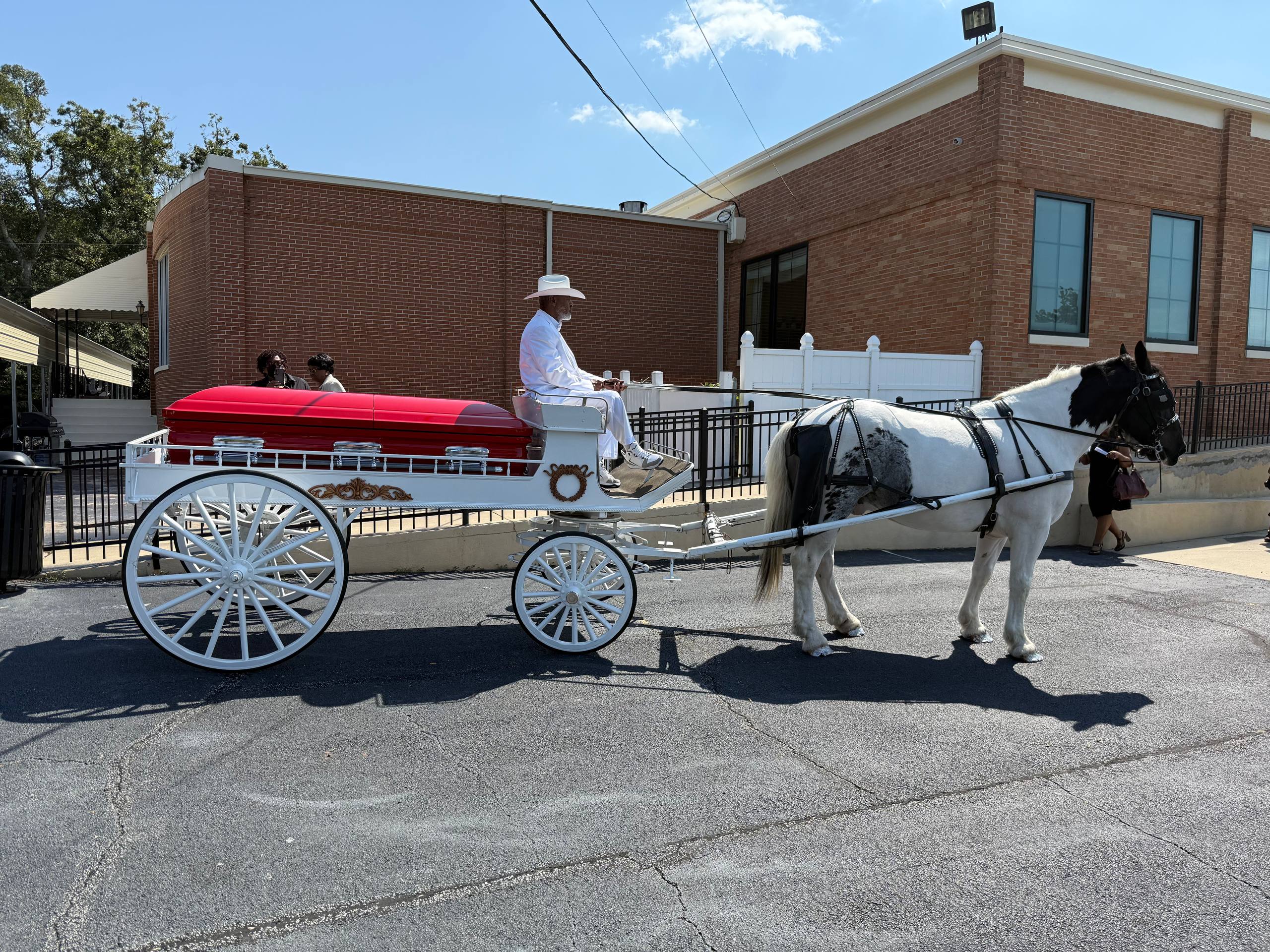 White horse pulling funeral carriage with red casket leaving a brick chapel