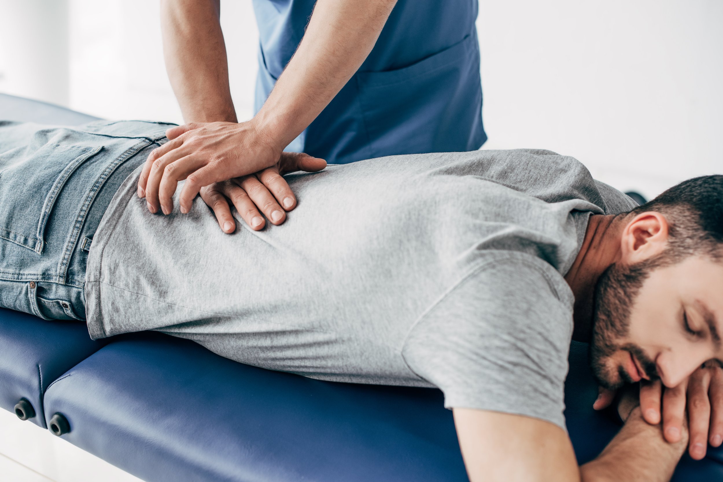 A man receiving chiropractic treatment on his lower back while lying face down on a treatment table, with a healthcare professional applying pressure with both hands.