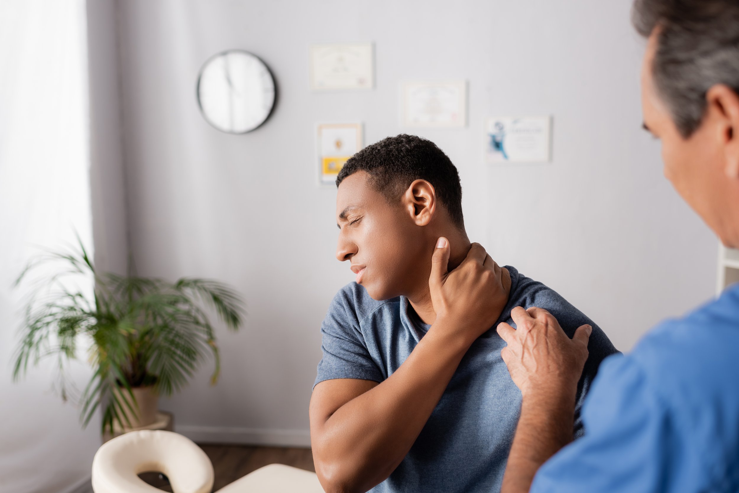 A person with curly hair experiencing neck pain while being examined by a healthcare professional in an office setting.