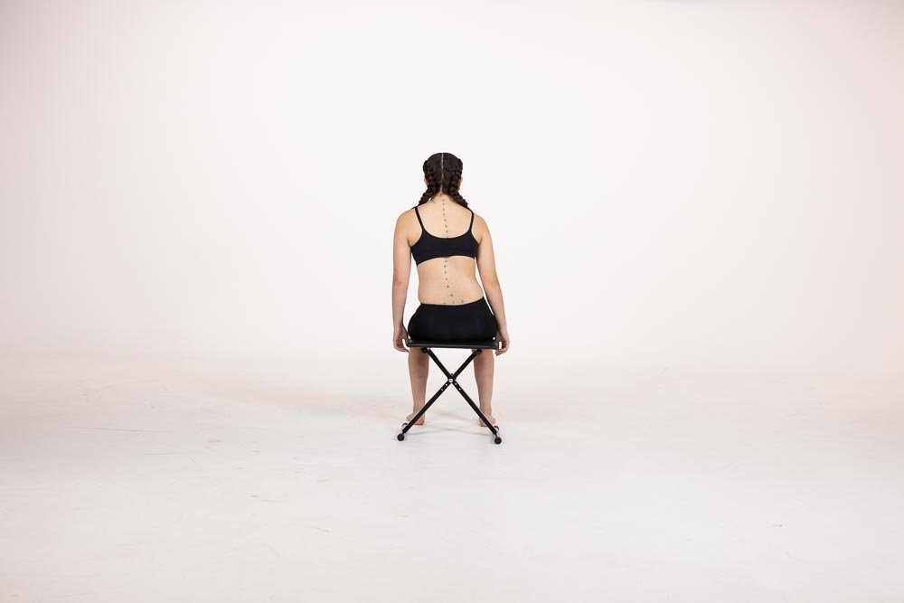 A woman with braided hair sitting on a stool with her back facing the camera, wearing a black sports bra and black shorts, in a plain white room.