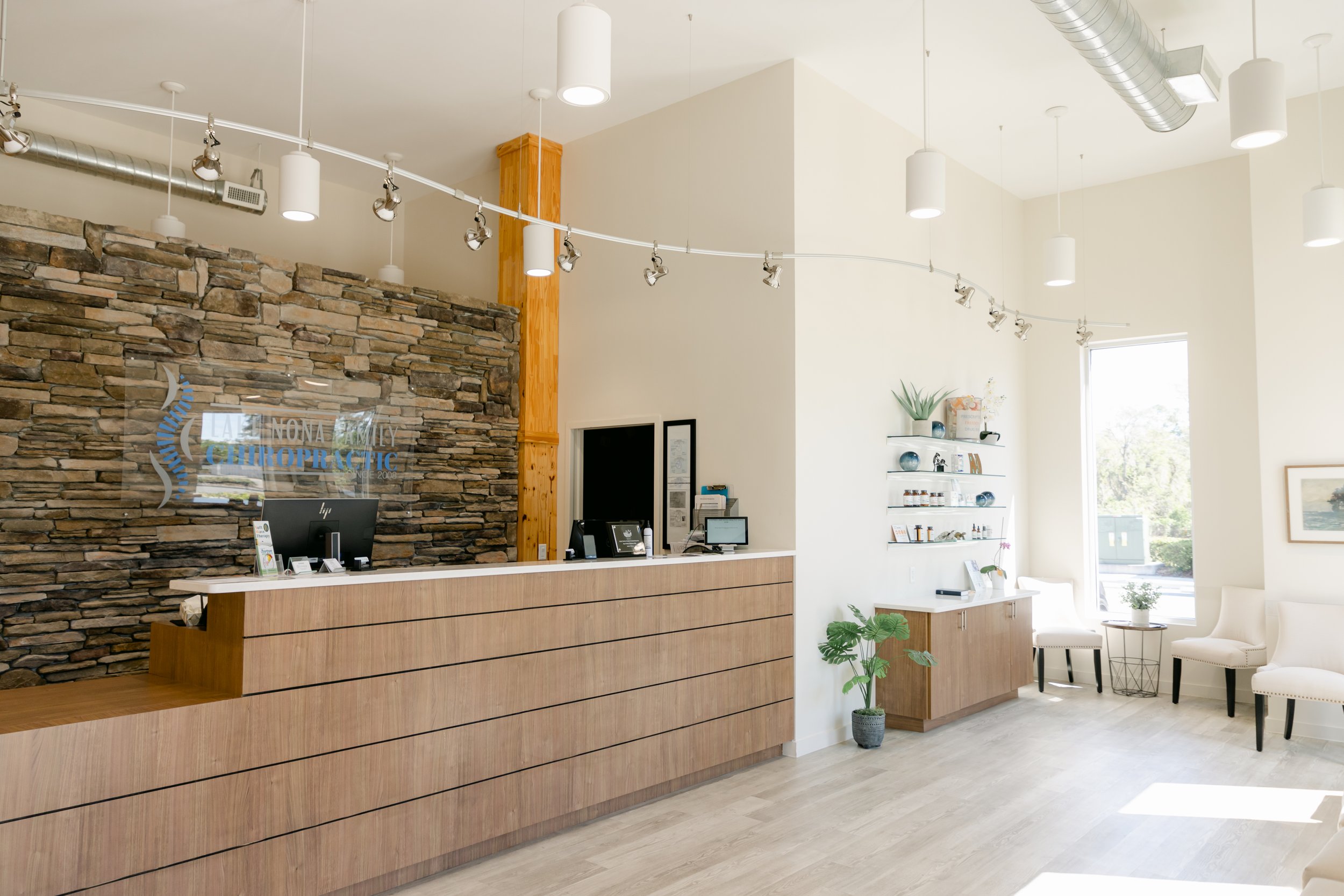 Bright and welcoming reception area featuring a stone accent wall with a logo, a wooden reception desk, and seating area with white chairs near large windows letting in natural light.