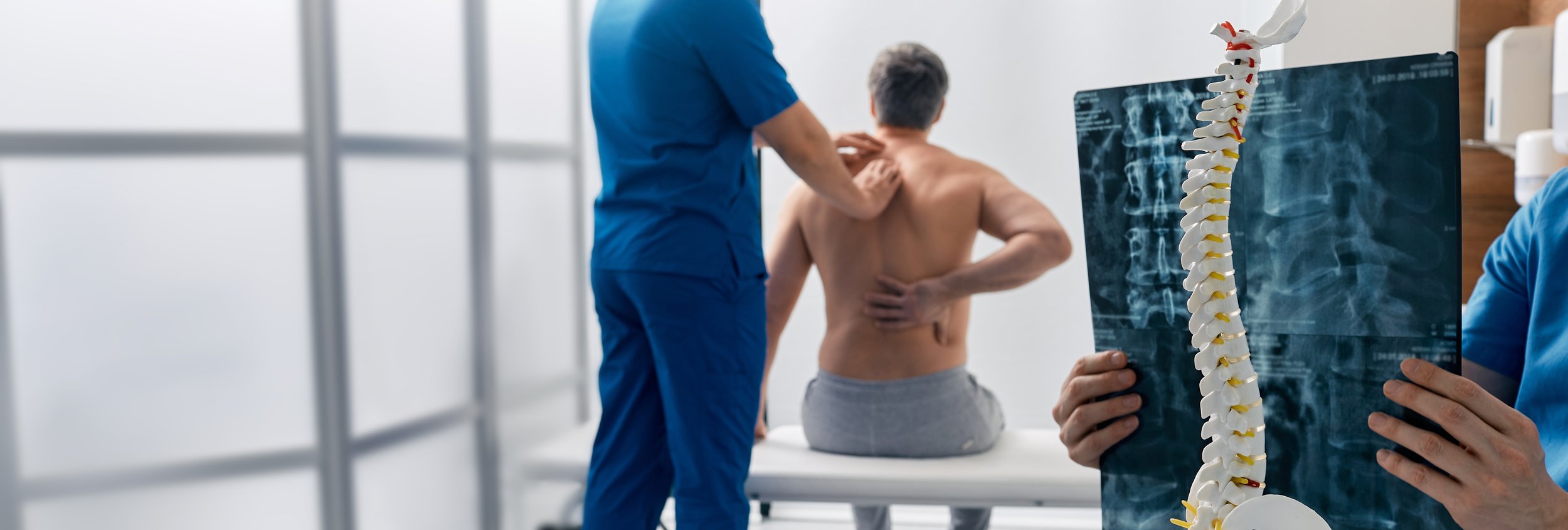 Doctor examining a patient's back while holding an X-ray image of a spine in a medical clinic.