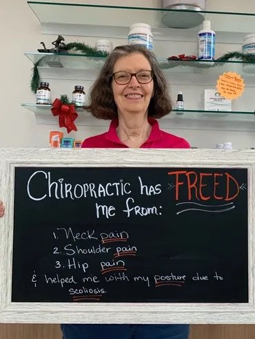 A smiling woman holding a chalkboard sign about relief from neck, shoulder, and hip pain and improved posture due to scoliosis
