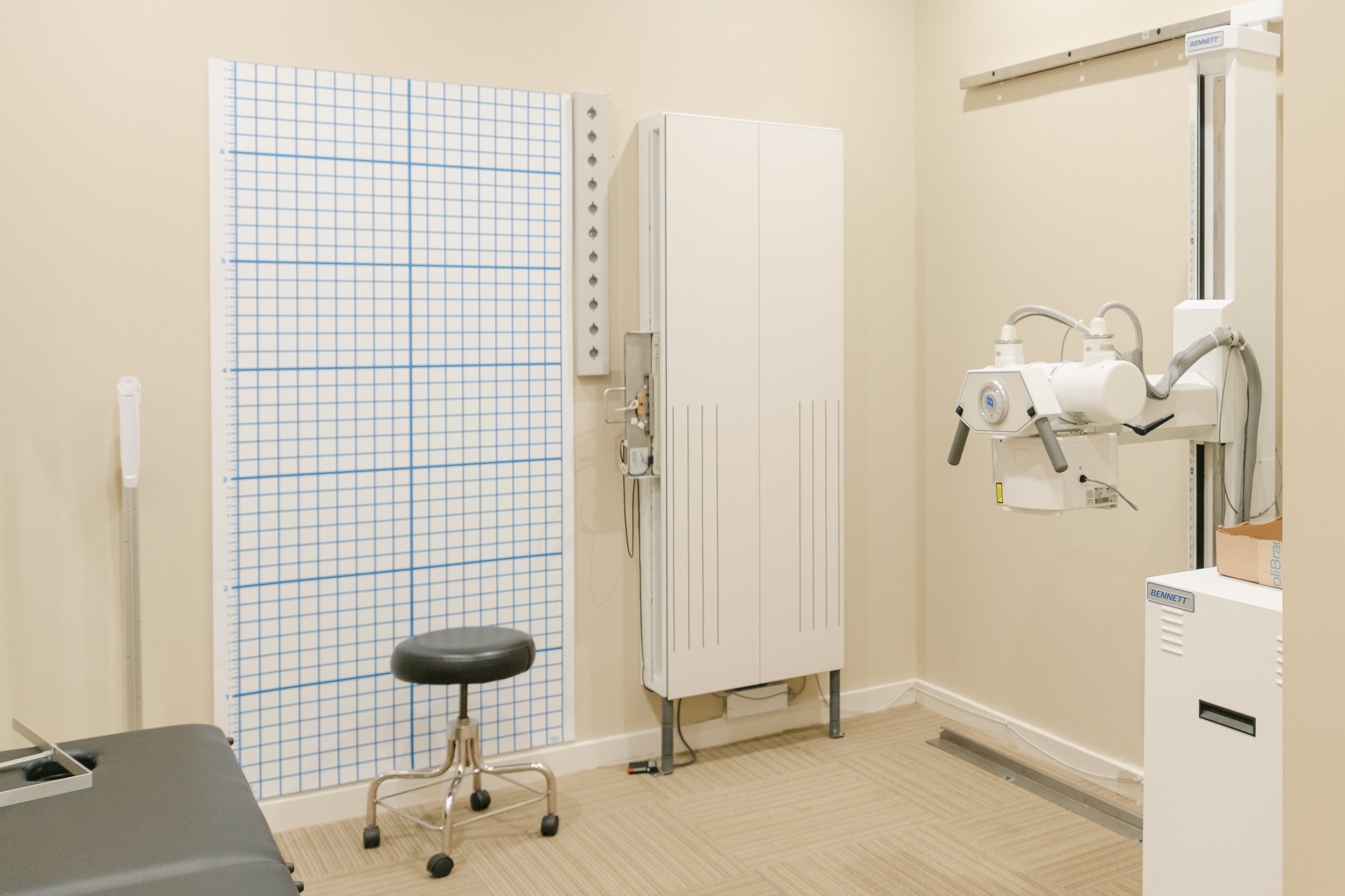 Medical examination room with radiology equipment, a grid chart on the wall, a stool, and medical cabinet.