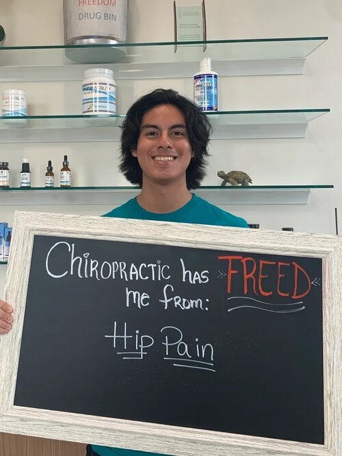 Person holding a chalkboard sign that says chiropractic has freed me from hip pain, smiling inside a room with shelves and medical supplies.