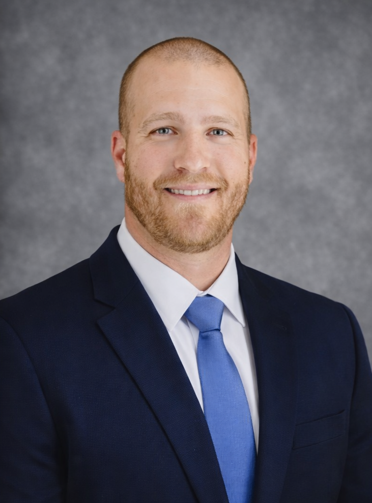 Headshot of Dr. Frank Berzanskis Jr., wearing a dark suit, white shirt, and blue tie, smiling against a gray background.