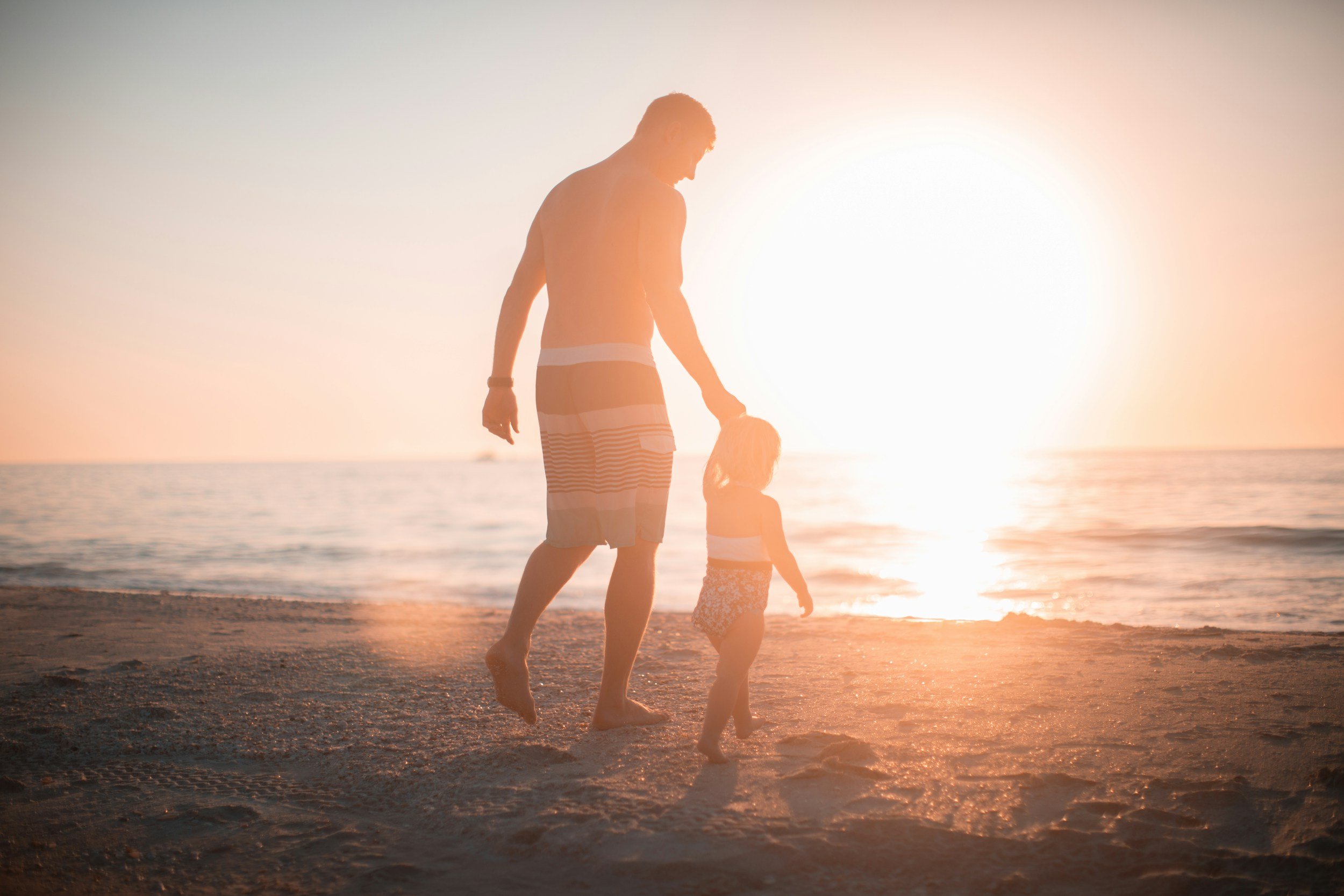 A silhouette of a person and a child walking on the beach at sunset, with the sun low on the horizon and the ocean in the background.