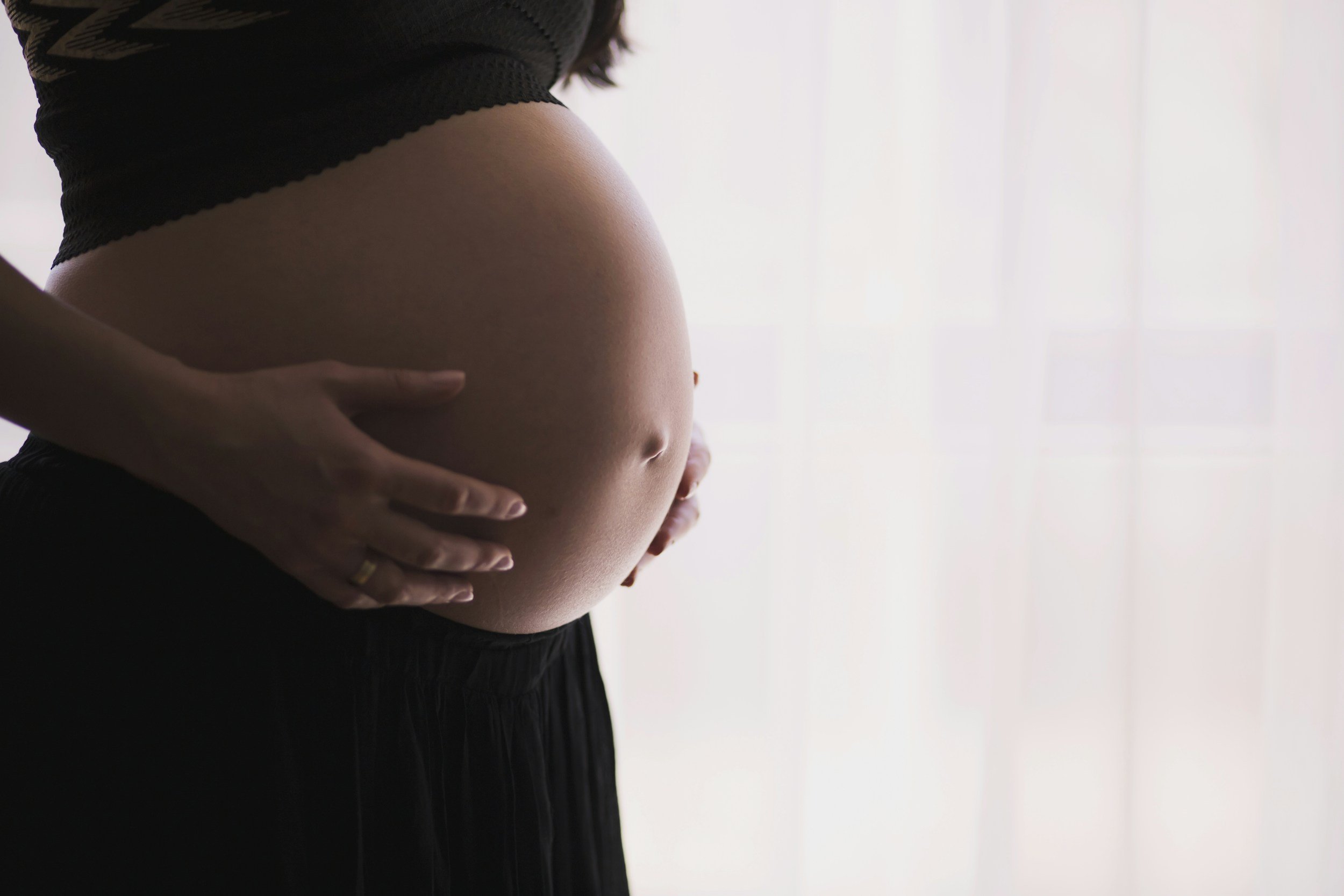 A pregnant woman cradling her belly with both hands, wearing a black top, standing near a bright window.