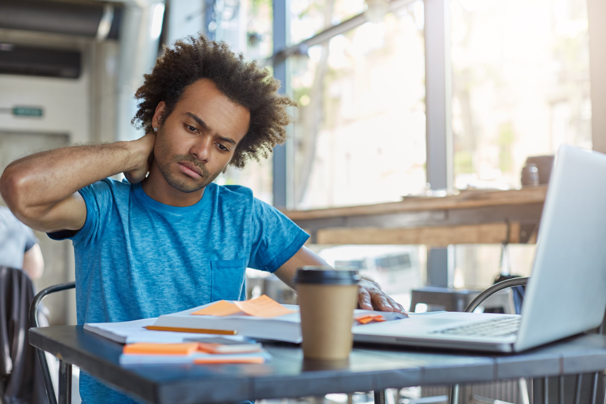 A man with curly hair wearing a blue t-shirt, sitting at a table in a coffee shop, looking at papers with a worried expression, with a laptop and a coffee cup on the table.