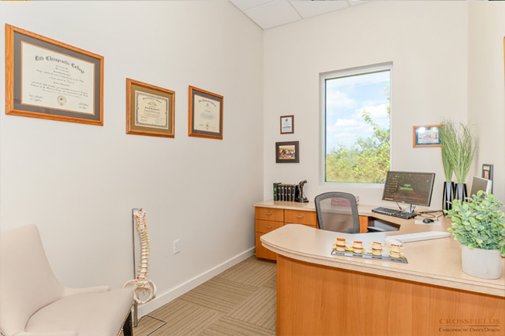 Office room with framed diplomas on the wall, a chair, a computer, a window showing trees outside, and a model of a human spine.