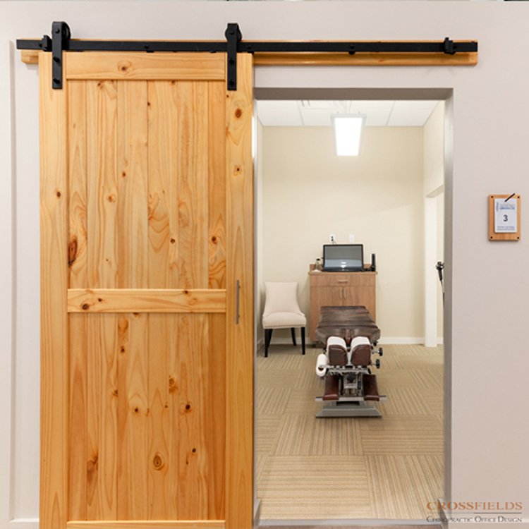 Hospital examination room with a wooden sliding barn door, a chair, a cabinet with a computer, and an examination table.