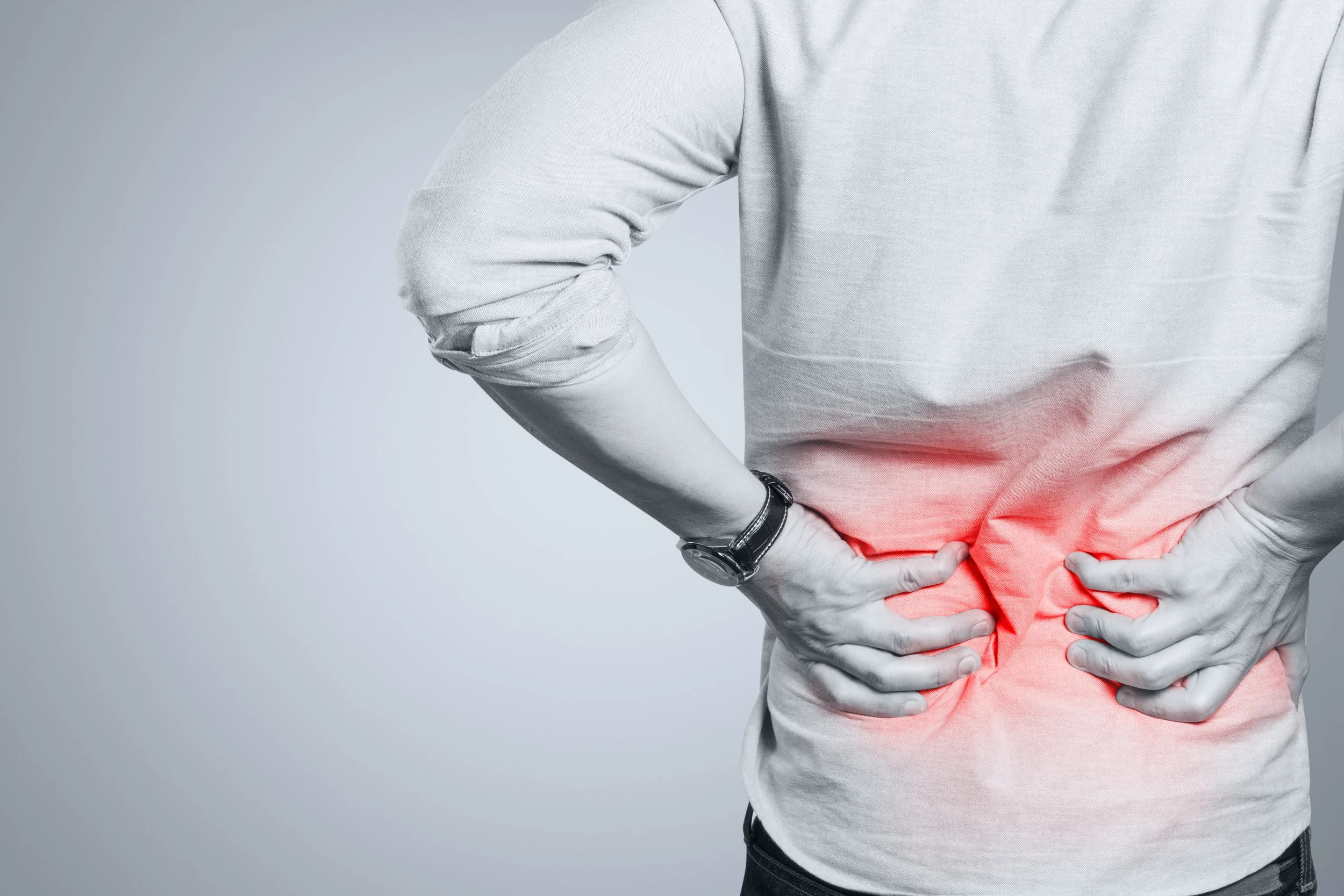A person wearing a gray shirt is holding their lower back with both hands, indicating pain or discomfort, with a red highlight on the lower back area.