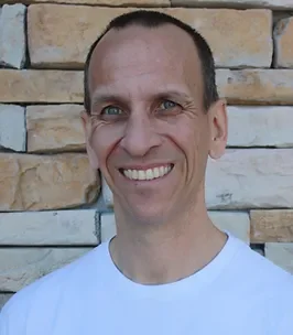 John Bruinsma, LMT, Massage Therapist, a smiling man with short brown hair and blue eyes, wearing a white t-shirt, standing in front of a stone wall.