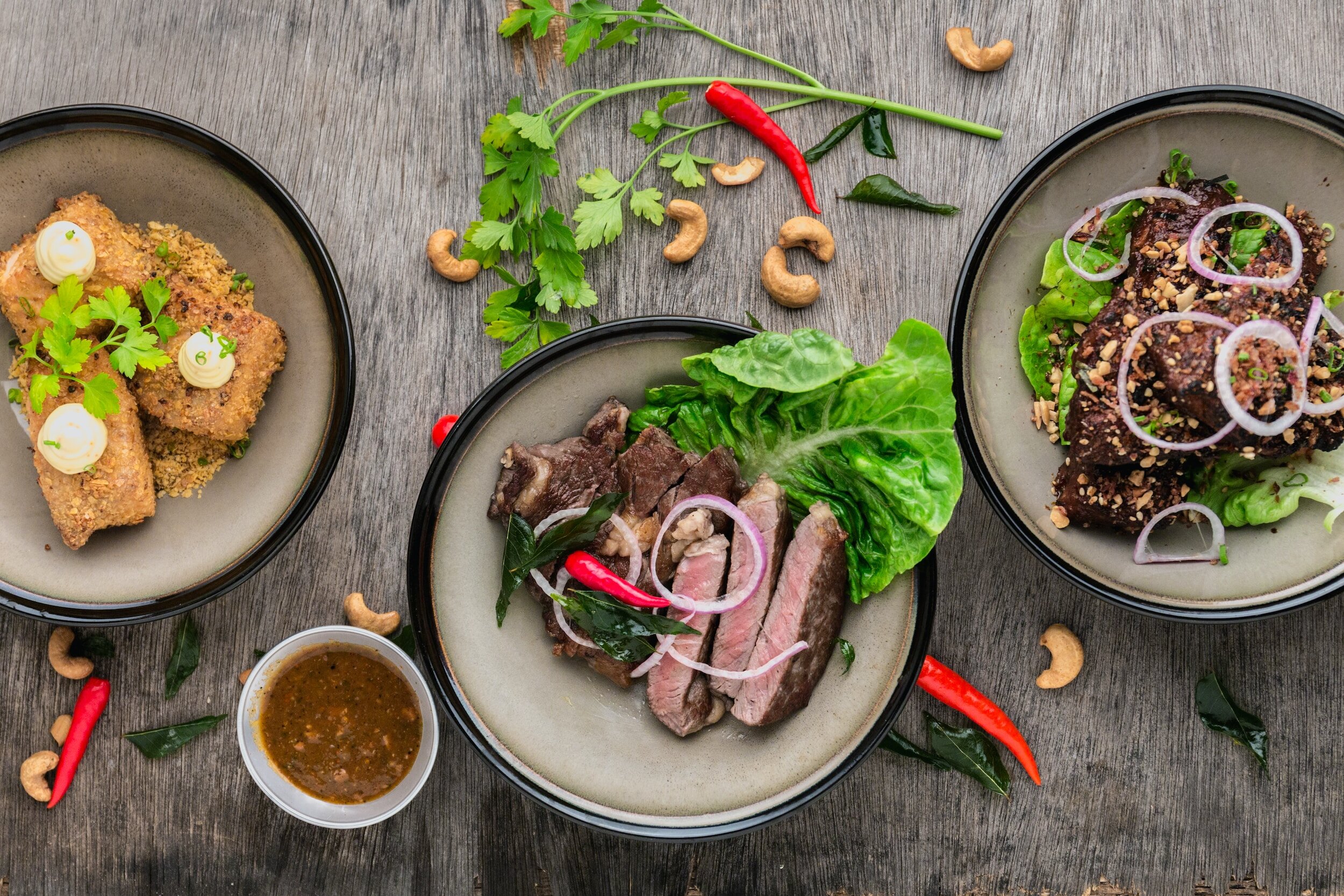 Three bowls of Asian-style dishes on a gray wooden table, surrounded by herbs, chilies, cashews, and a small dish of dipping sauce.