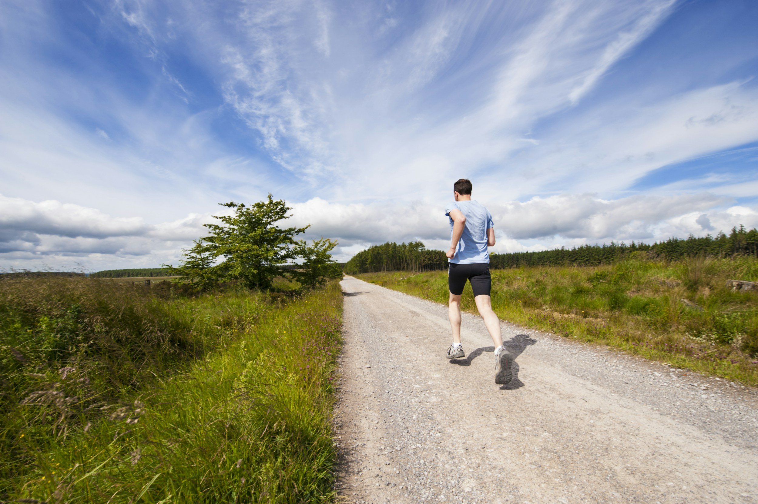 Man jogging on a dirt trail in a rural area with green grass and trees, under a blue sky with scattered clouds.