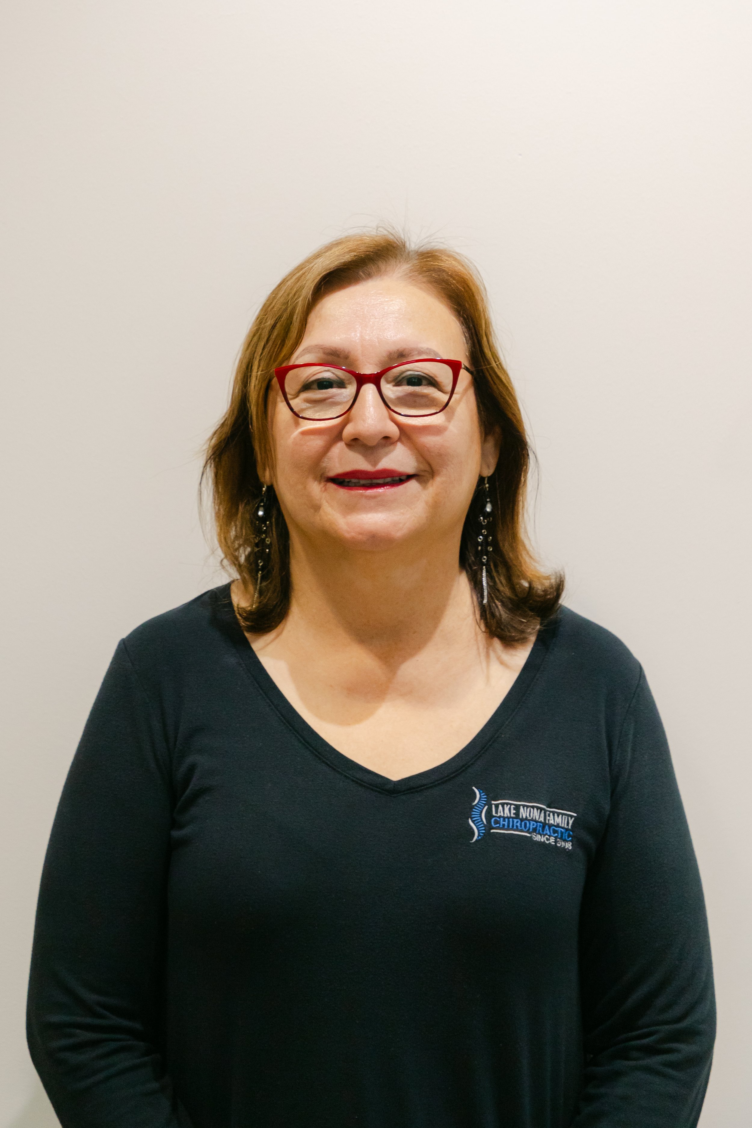 Blanca Balk, Patient Coordinator, with shoulder-length reddish-brown hair and red glasses, wearing a black long-sleeve shirt with a Lake Nona Chiropractic logo.