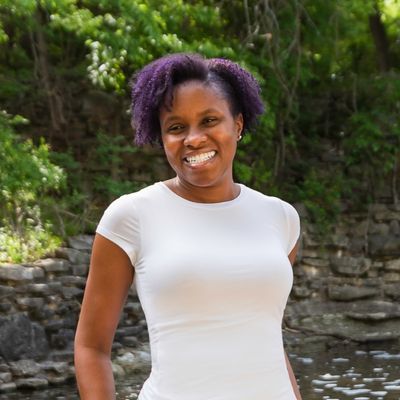 Merissa Si-Lence smiling outdoors in a white shirt with purple natural hair, standing near a creek