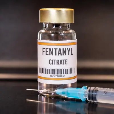 Pharmaceutical fentanyl citrate glass injection vial with gold metal cap and white label, alongside a blue clinical syringe on a dark reflective surface — educational image by Medivira, fentanyl addiction treatment clinic in Collin County, Texas.