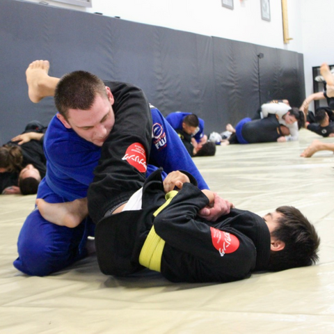 Alt Image Text: A skilled Jiu-Jitsu Fundamental teacher, wearing a blue belt, demonstrates a technique as he is placed in a controlled submission hold, specifically an armbar, by his focused student in a black gi during a training session at the Renzo Gracie of Weston gym, where they consistently practice and improve their grappling skills under the guidance of experienced instructors.