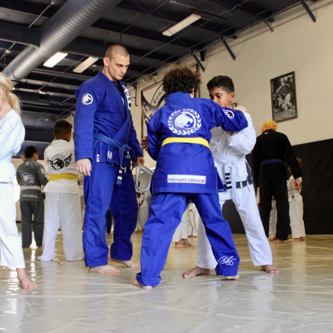 Two enthusiastic kids practicing jiu-jitsu fundamentals on the pristine blue mats at the Renzo Gracie of Weston Gym, closely supervised by experienced black belt owner Adrian Alsina, ensuring their safety and proper development of martial arts skills in a vibrant and welcoming environment.