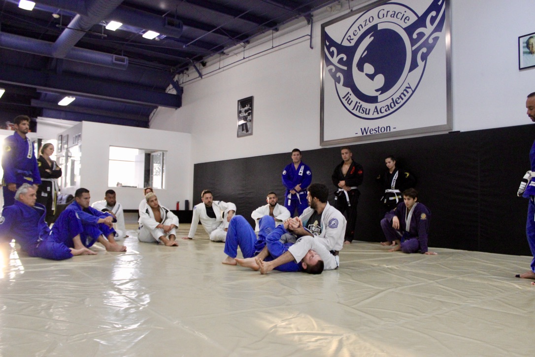 Adrian Alsina, a skilled black belt and the current owner of Renzo Gracie of Weston Gym, expertly demonstrates a complex jiu-jitsu submission hold on an engaged adult male student during a men's class, highlighting the finesse and technique required for this intricate martial art in the modern, well-equipped Renzo Gracie of Weston training facility.