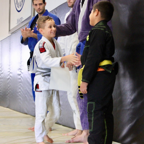 Two young jiu-jitsu athletes displaying exceptional sportsmanship, shaking hands with genuine smiles on their faces, standing side by side after being awarded medals at a competition, signifying their dedication and respect for each other's skills and efforts.