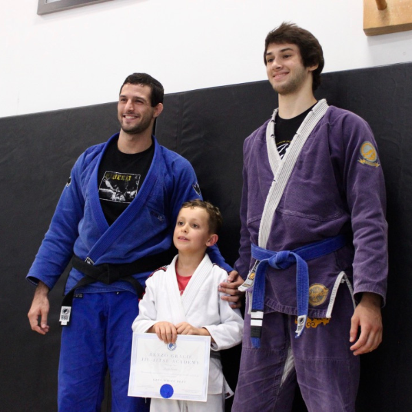 A young martial arts student beaming with pride as he receives his award for successfully advancing to the next belt level, stands between two esteemed instructors of the school - one of whom is the dedicated and accomplished head coach, Adrian Alsina. They pose together, as the instructors hold up the newly earned belt, showcasing the fruits of the student's hard work and commitment to his craft.