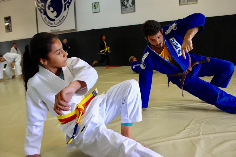 Head Coach Adrian Alsina, wearing his then-brown belt, skillfully demonstrating a Jiu-Jitsu technique to a group of focused teenagers, all wearing Gi uniforms, at the renowned Renzo Gracie Jiu-Jitsu Academy of Weston, with the academy's logo and multiple black belts hanging prominently in the background.