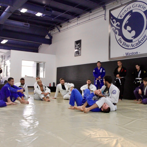 Adrian Alsina, an experienced instructor at the renowned Renzo Gracie Jiu-Jitsu Academy of Weston, expertly demonstrates a submission hold to an attentive group of adult beginners in a well-equipped training facility. Wearing a traditional Brazilian Jiu-Jitsu gi, Adrian skillfully guides his training partner into the hold, highlighting the importance of leverage and technique for successful self-defense. Fellow students eagerly observe and absorb the knowledge, eager to practice and refine their own grappling skills under Adrian's watchful eye.