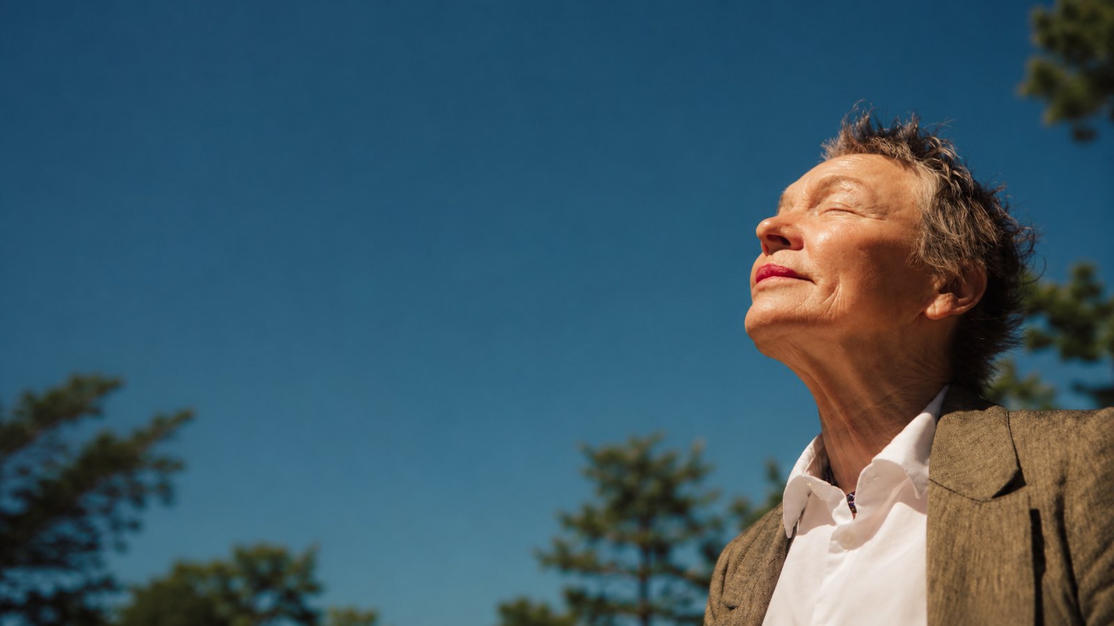 An older woman outdoors with her face turned up to the sun against a clear blue sky.