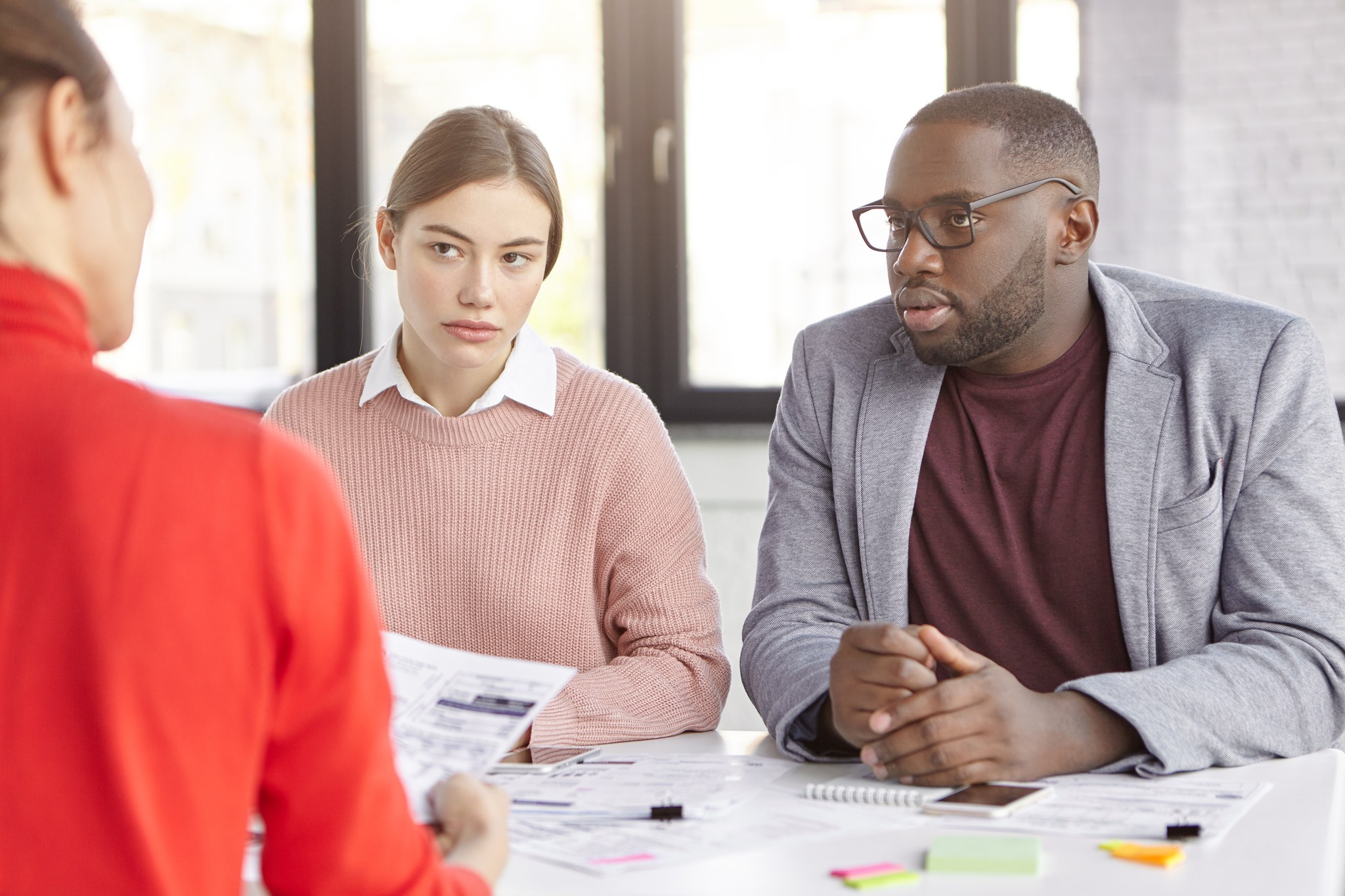 Two colleagues at a table in a quiet office, one still speaking while the other has looked down and away, the moment a message stops landing