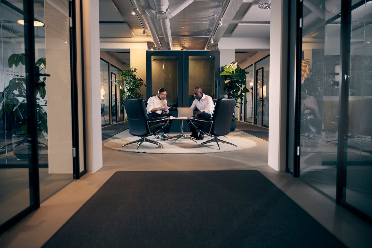 Two colleagues working quietly across a table in a modern glass office, the work continuing the same way it always has