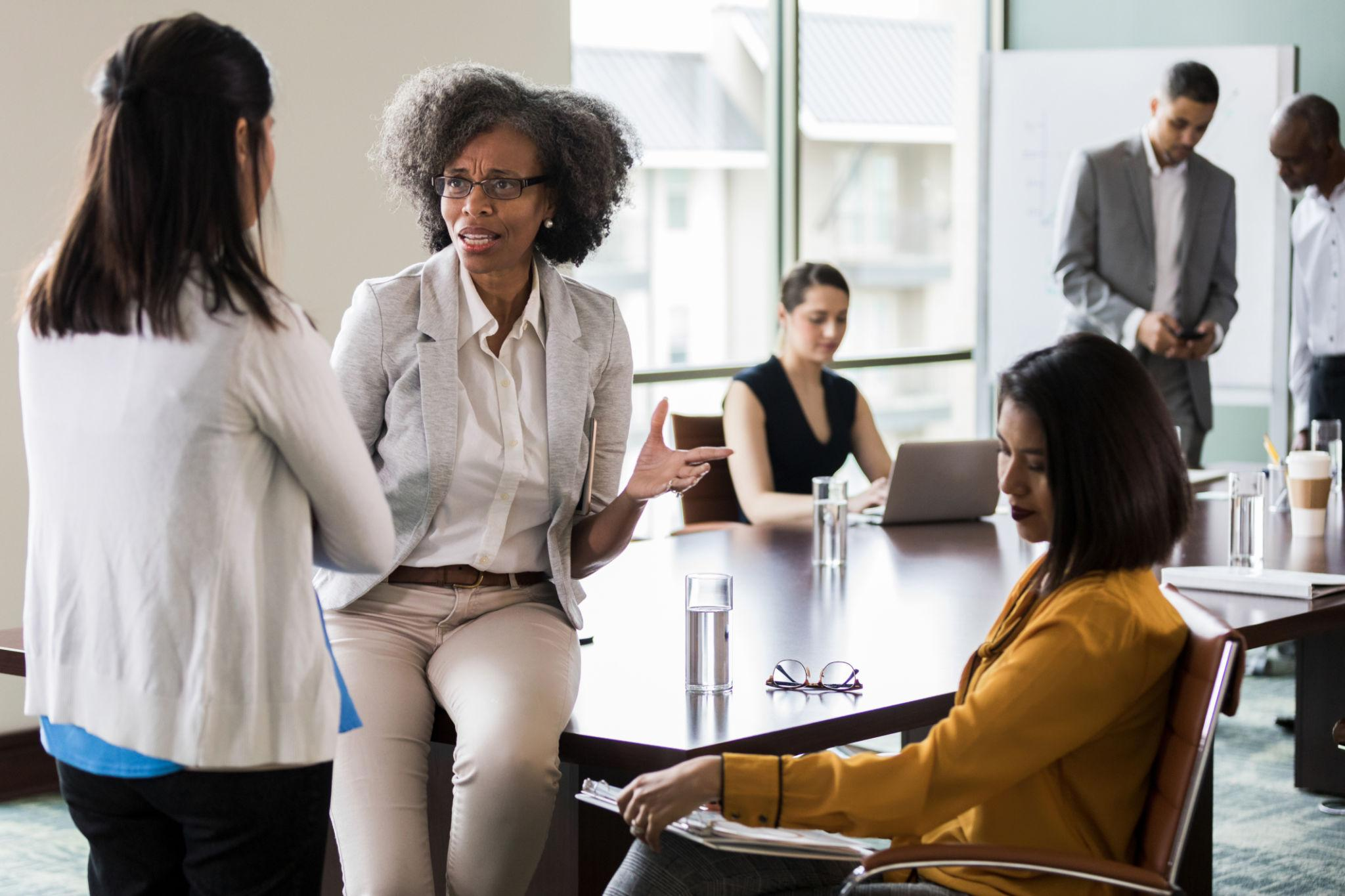 A senior leader mid-conversation with a colleague after a meeting, others still in the room, IEXDG leadership culture