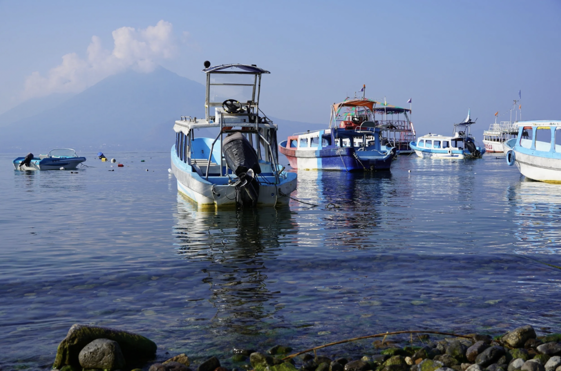 Boats are moored in the crystal clear waters of Lake Atitlan in Guatemala on a sunny day with volcanos in the background.