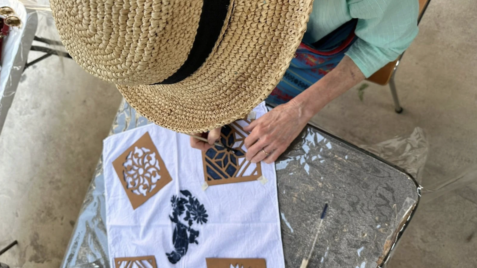 A woman in a hat is bent over and drawing with a stencil onto fabric during a natural indigo dye workshop in Guatemala